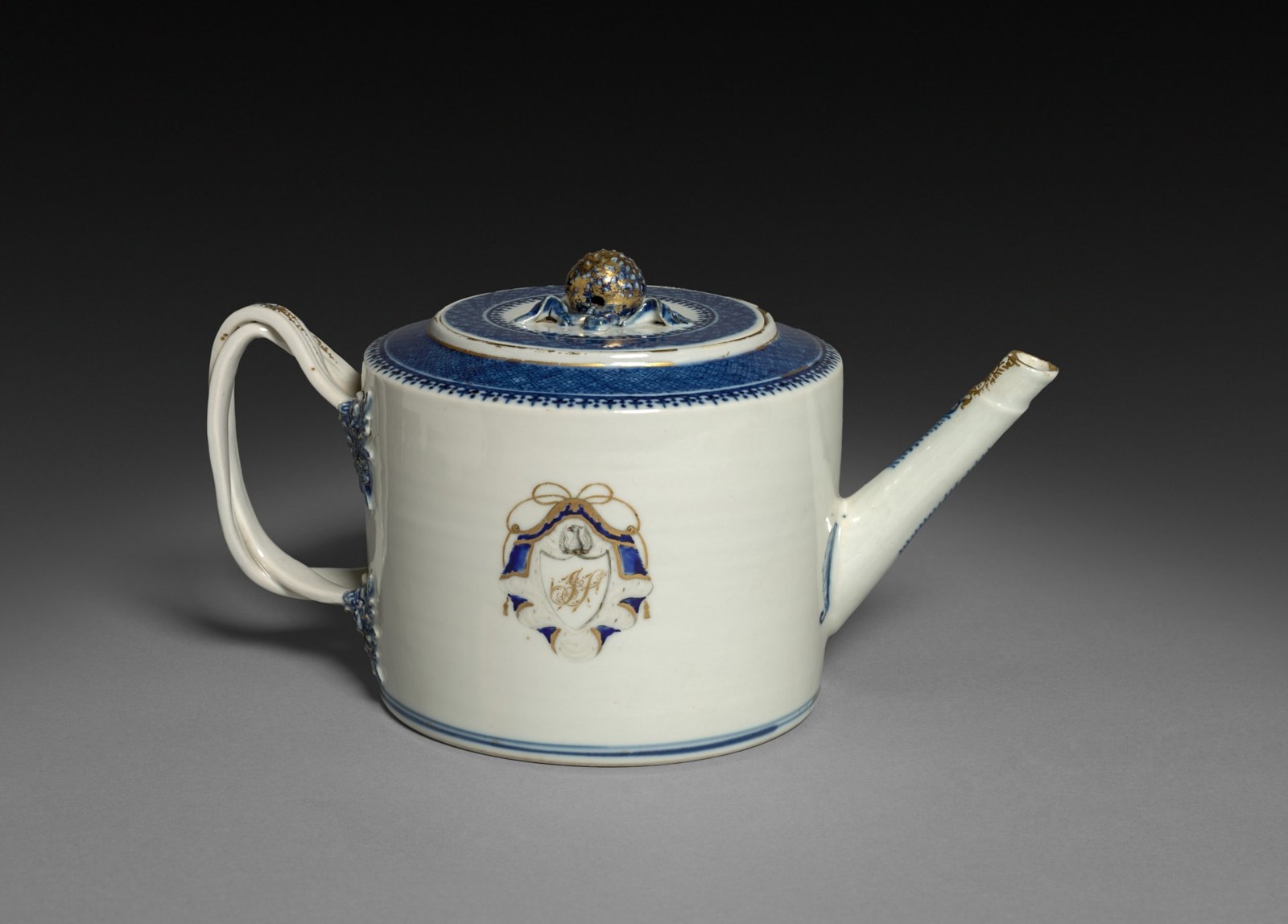 A cylindrical white teapot carries a blue-and-gold shield-shaped cartouche containing intertwined letters. A small ornament sits above, while blue patterns encircle the lid and upper body.