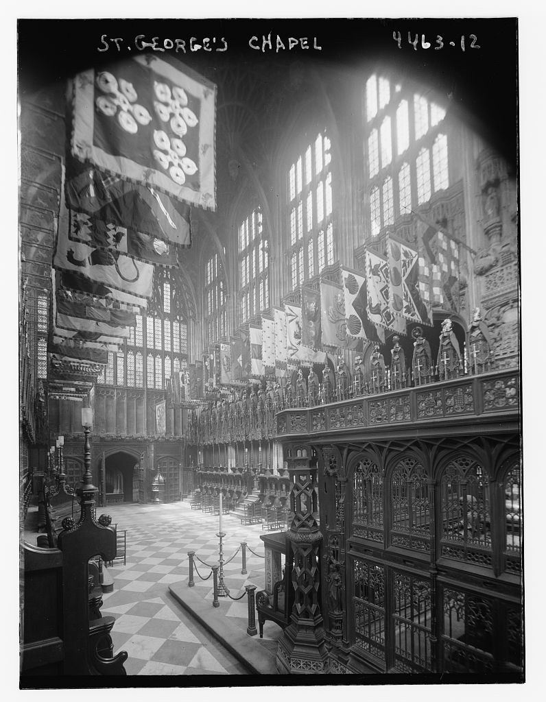 Rows of banners hang over carved stalls. Helms and crest-like ornaments stand along the canopies, while small rectangular plates cover parts of the woodwork below.