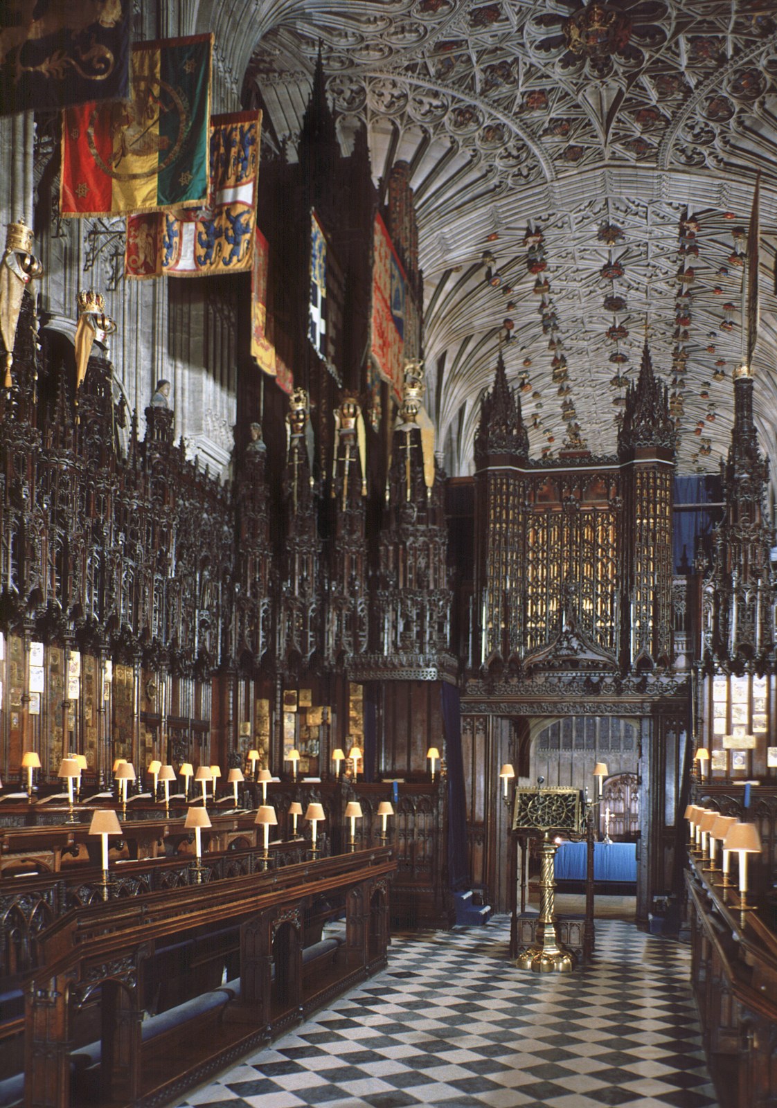 Large coloured banners occupy the upper left, with helms and ornaments over the stalls. Shields also appear within the patterned vault, while lamps, seats, and a patterned floor organize the occupied room.
