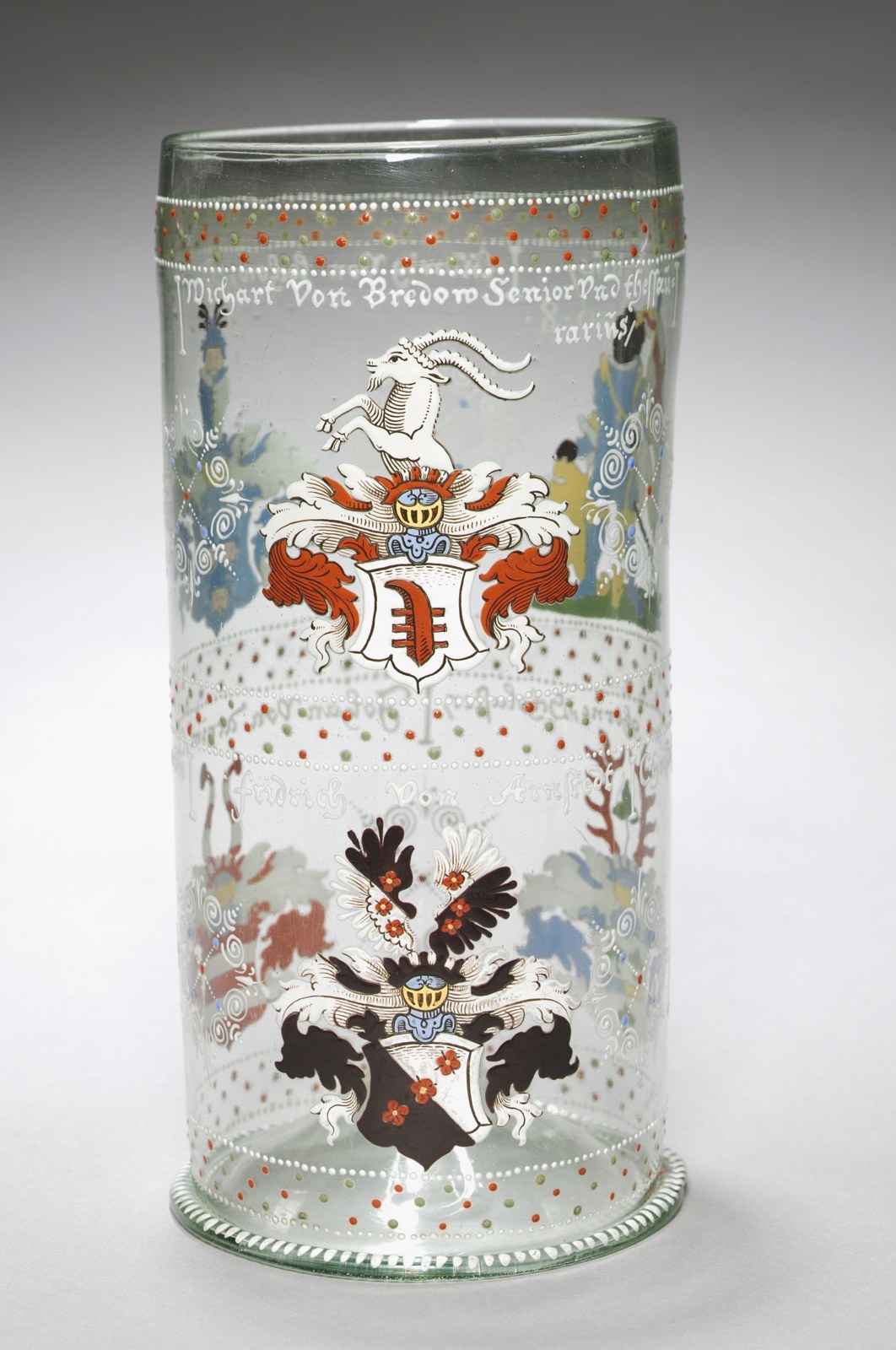 A tall cylindrical beaker carries enameled achievements, lettering, dotted bands, and figures. The curved glass turns part of the display away from the camera.