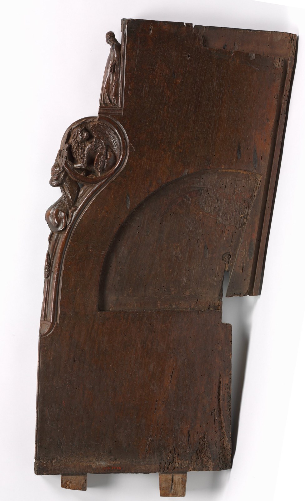 A dark wooden panel has an irregular curved profile and projecting joints. Carved figures and animal-like forms occupy part of the edge, while much of the broad face remains plain.