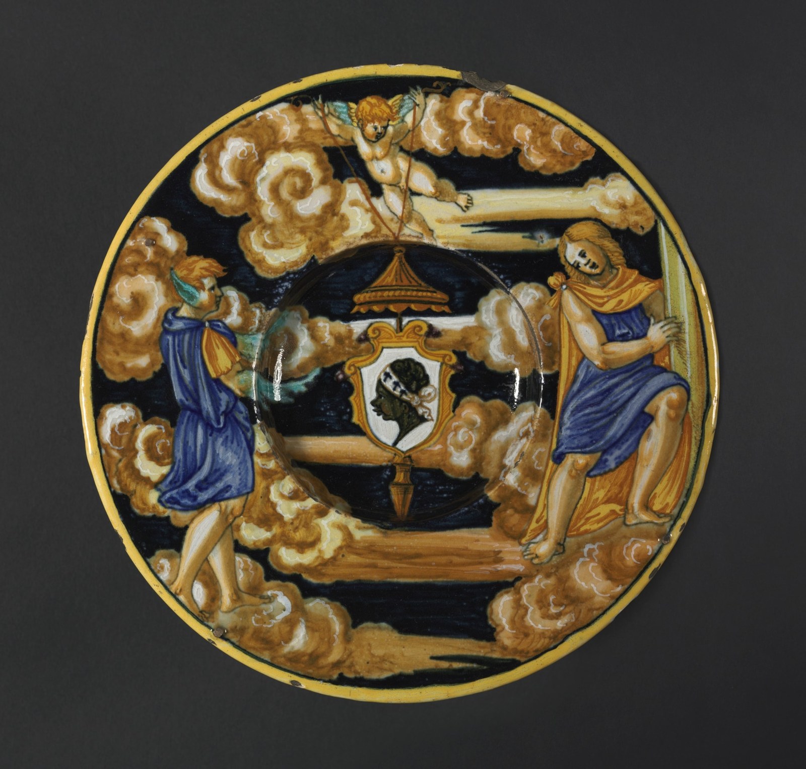 A small shield with a dark profile head sits near the centre of a richly painted plate. A winged child holds it amid larger figures, clouds, a column, and a yellow rim.